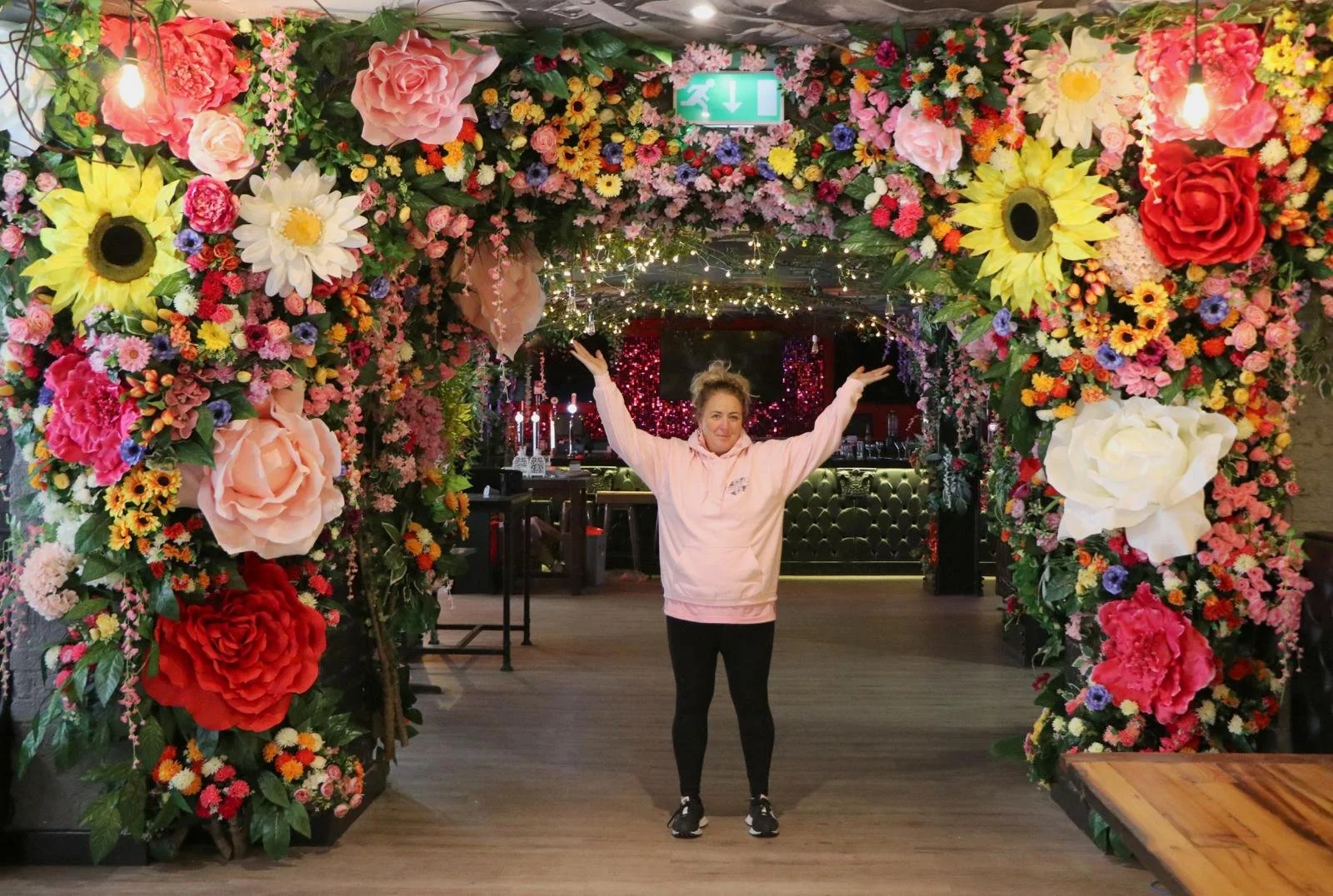 A woman standing under a large floral arch with her arms raised in a decorated indoor space.