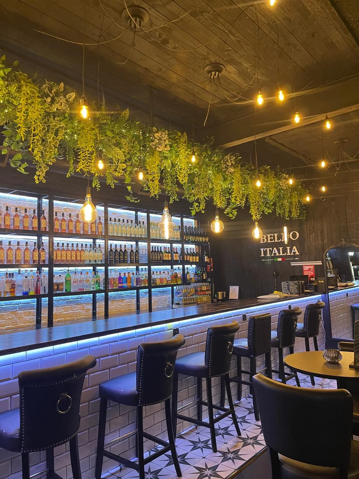 Interior of a modern bar with a row of black barstools along a white tiled bar counter illuminated by blue lights. Behind the bar are shelves filled with bottles, illuminated with cool lighting. Above the bar is green hanging foliage and warm Edison-