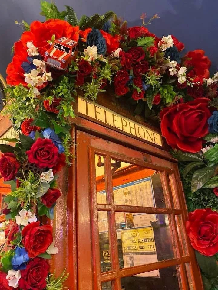 Decorative red telephone booth with a floral arrangement of red roses, white flowers, and greenery,on top, including a small toy car, in an indoor setting.