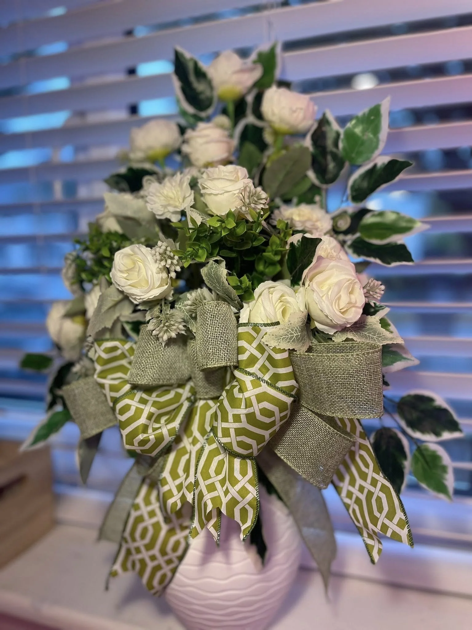 A bouquet of white roses, carnations, and greenery with a green and beige patterned ribbon in a pink vase