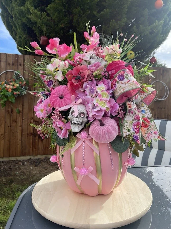 Pink pumpkin-shaped floral arrangement with pink pumpkins, skulls, flowers, ribbons, and decorative beads, placed on a wooden platform outside.