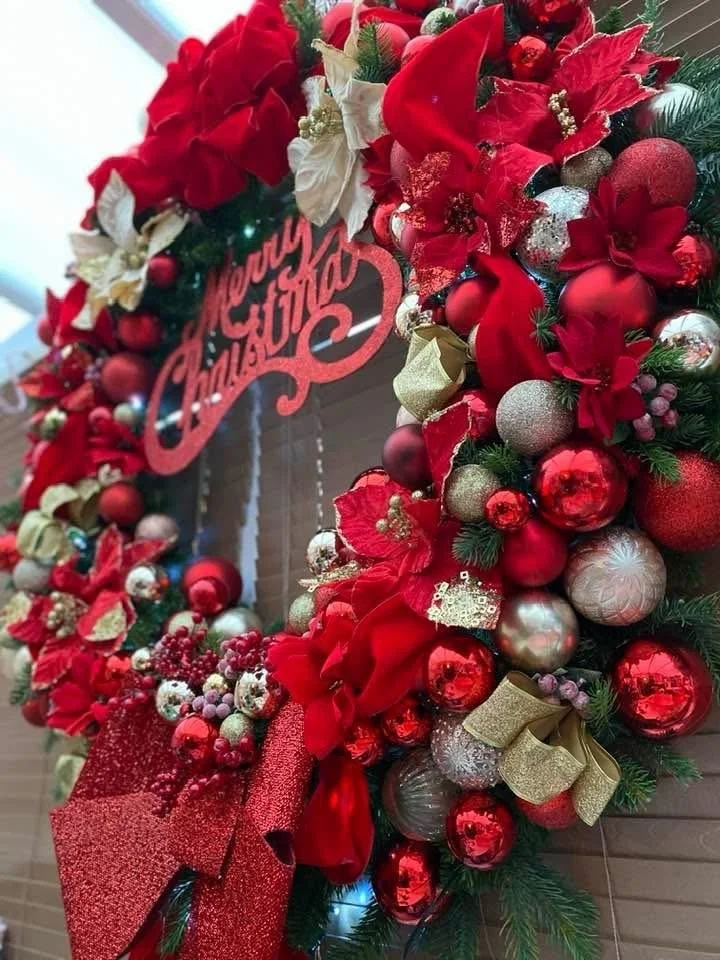 A Christmas wreath decorated with red and gold ornaments, bows, and flowers, with a sign that says 'Merry Christmas' in red lettering.