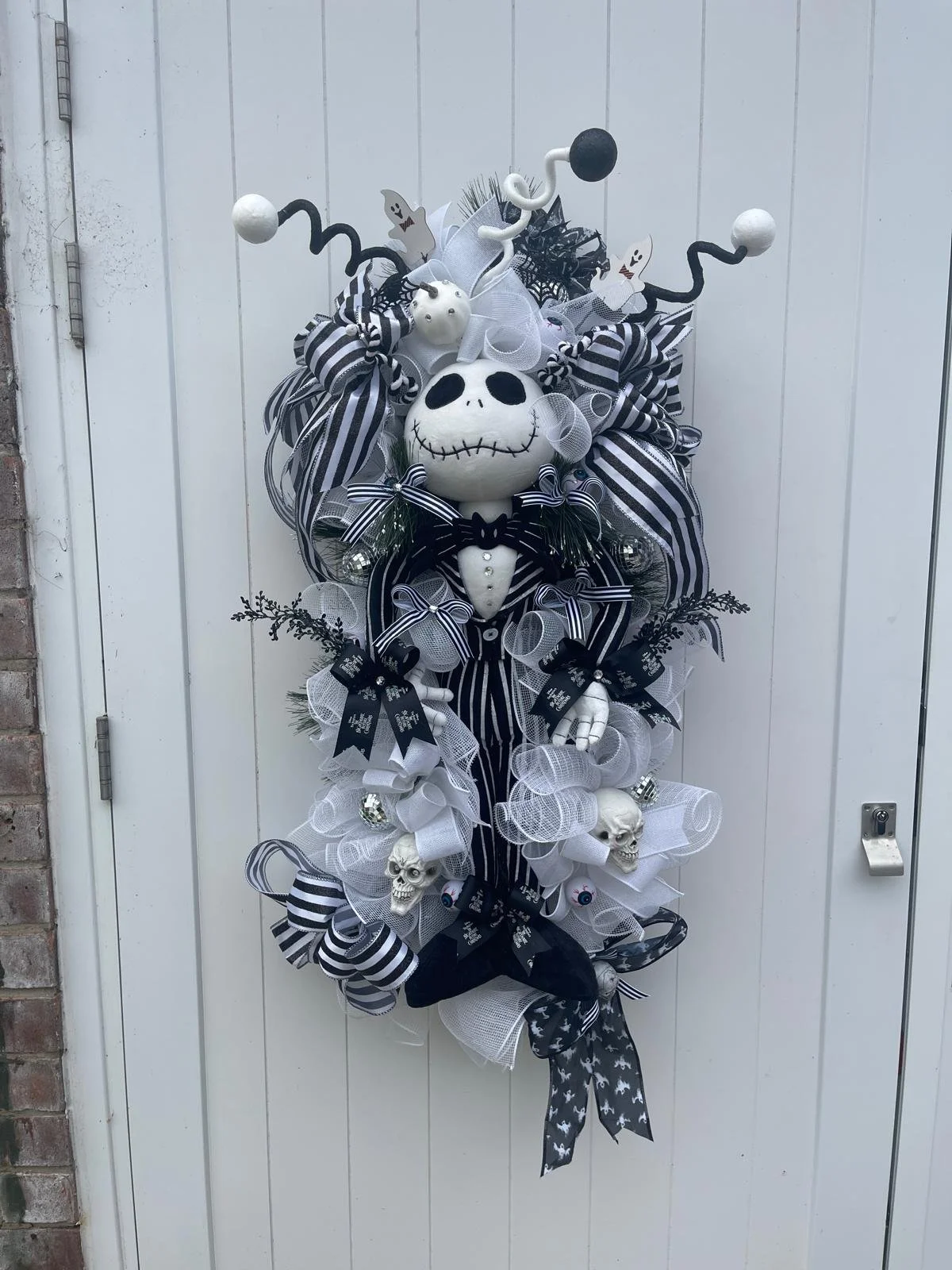 Halloween-themed wreath featuring a white skull with black eye sockets and stitched mouth, dressed in a tuxedo with a bowtie, surrounded by black and white ribbons, skulls, ghosts, spiders, and decorative elements in black, white, and gray colors.