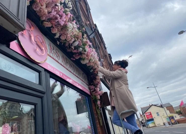 A woman decorating a storefront with pink and white flowers hanging from the awning, standing on a stool, in a small town with cloudy skies.
