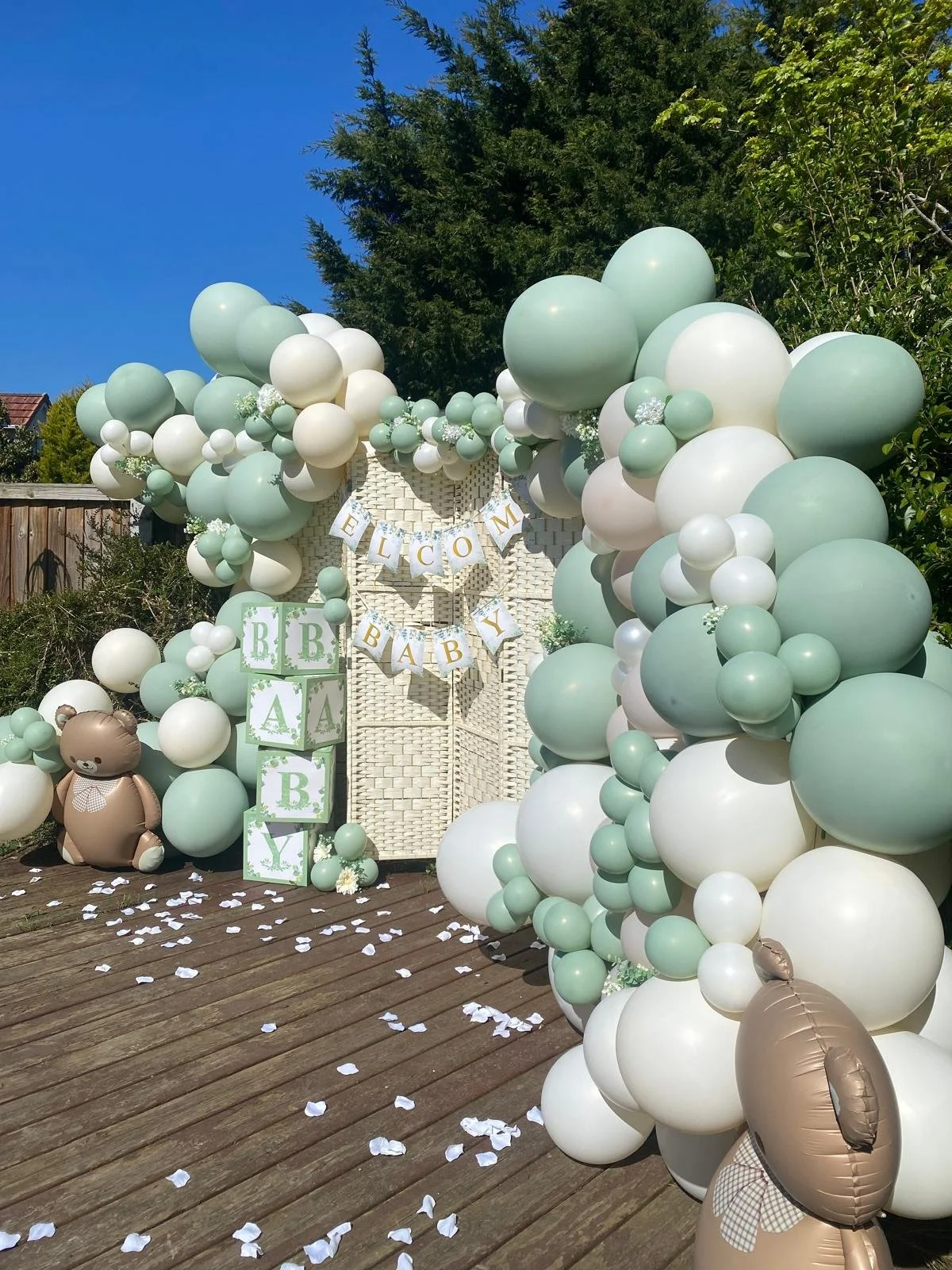 Decorative outdoor welcome setup with pastel green and white balloons, teddy bear balloons, and banners reading 'Welcome Baby'.
