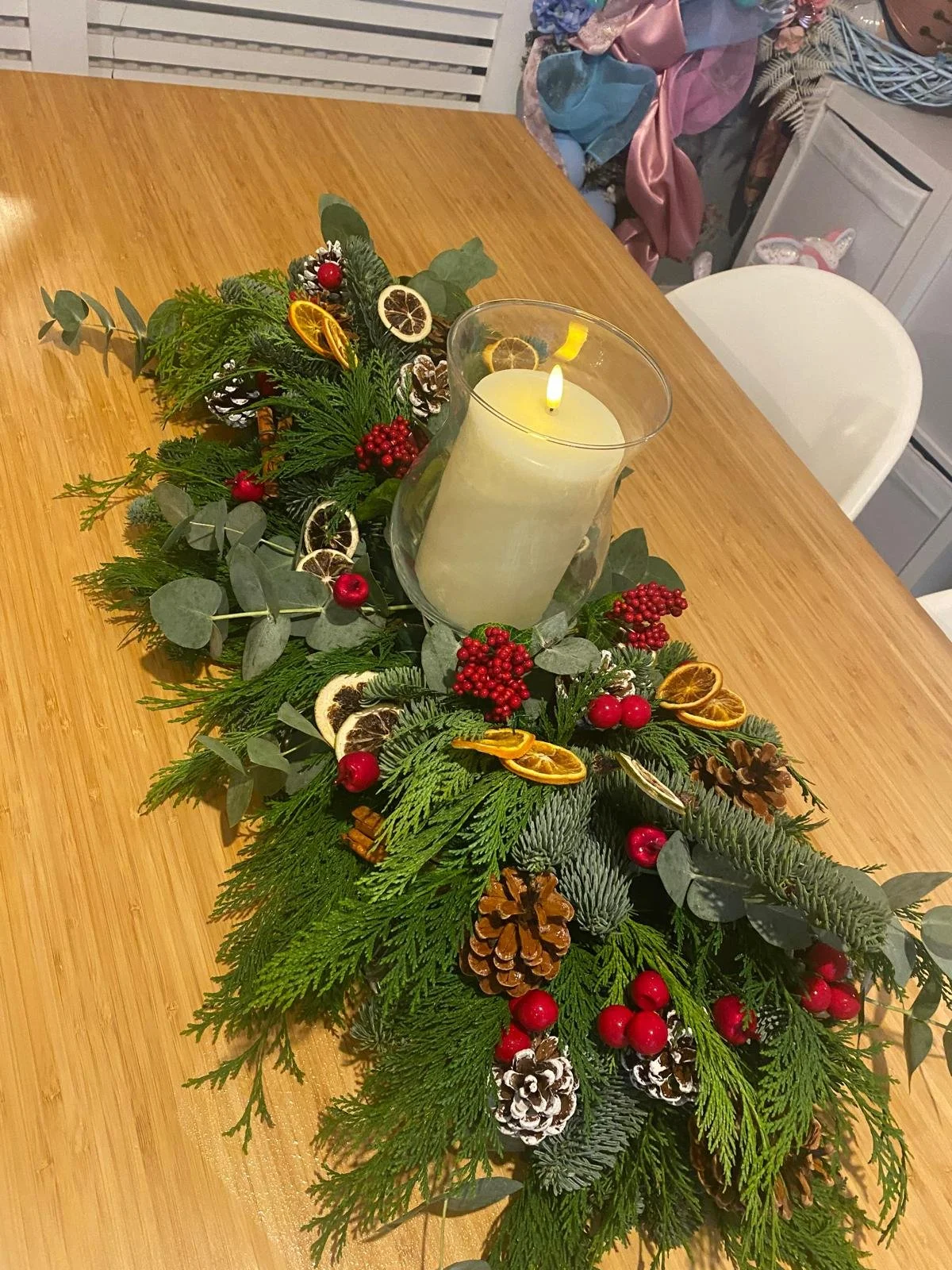 Festive holiday table centerpiece with evergreen branches, pinecones, dried orange slices, red berries, and a white candle in the center.