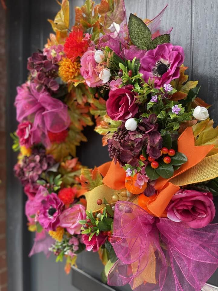 A colorful autumn wreath decorated with pink, orange, yellow, and purple flowers, complemented by red berries, green leaves, and pink mesh ribbons on a dark background.