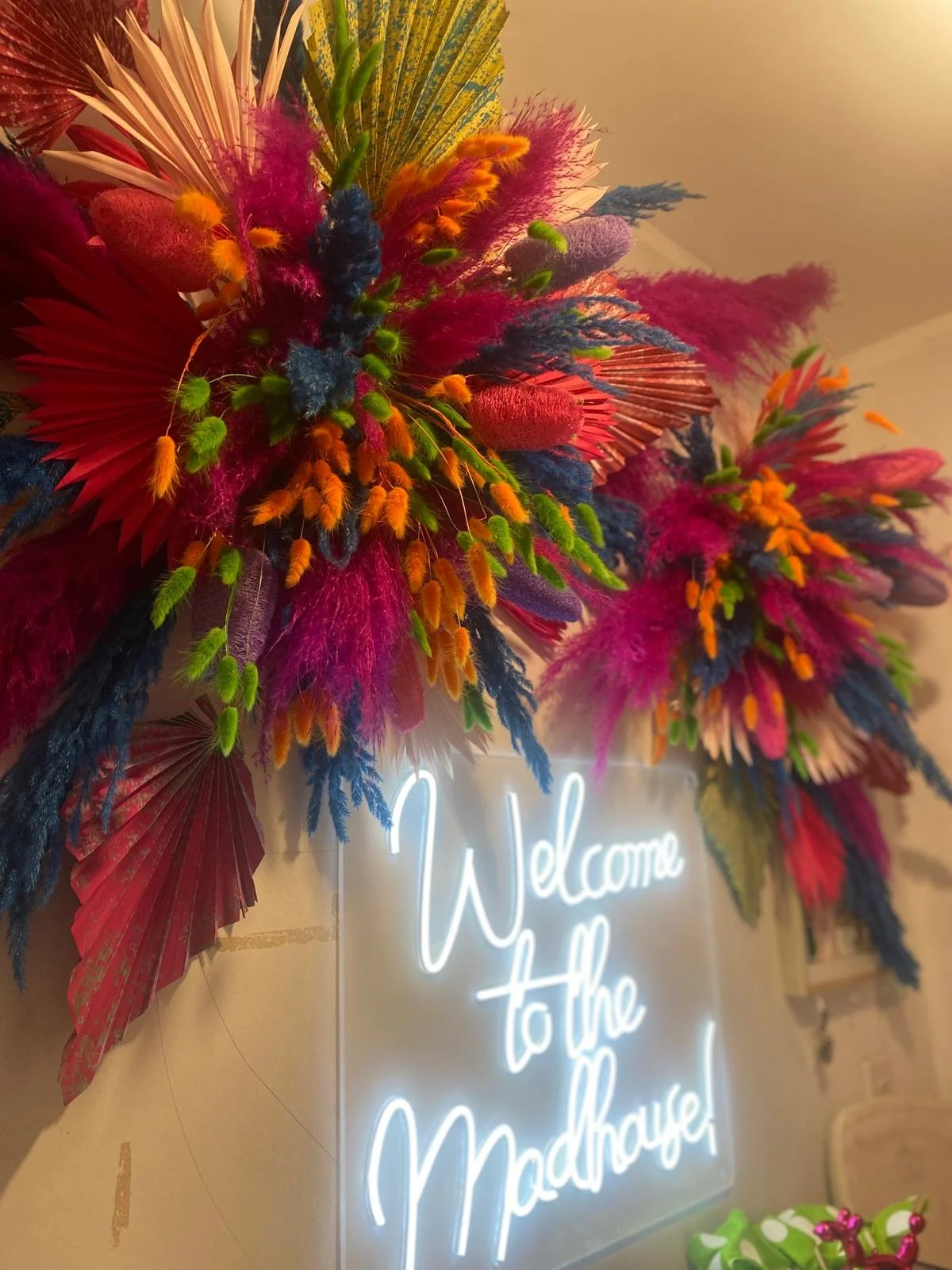 Colorful floral arrangement with pink, green, orange, blue, and red elements, and a neon sign that reads "Welcome to the Madhouse".