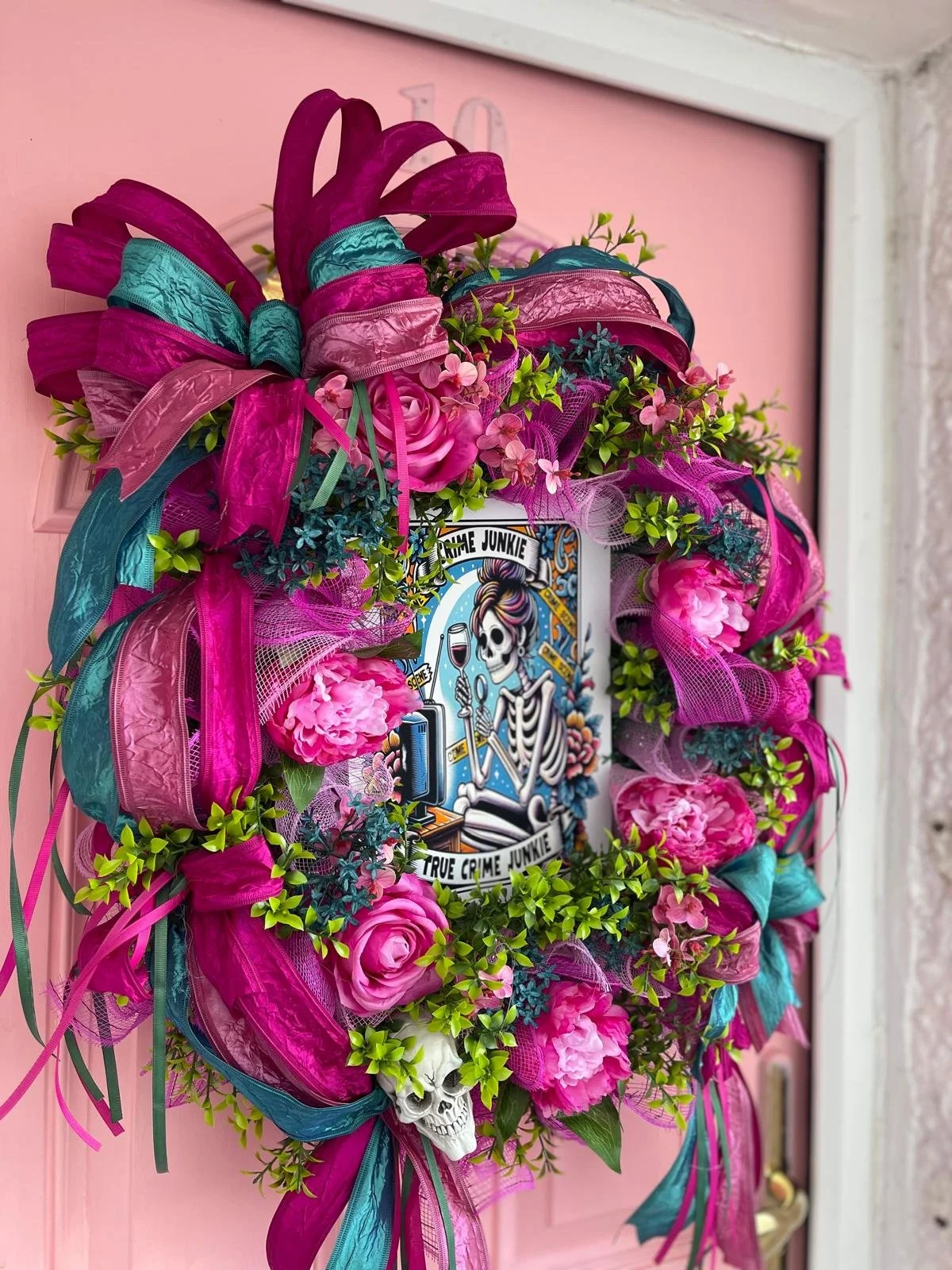 Colorful wreath with pink and teal ribbons, pink flowers, green leaves, and a small skull at the bottom, hanging on a pink door.