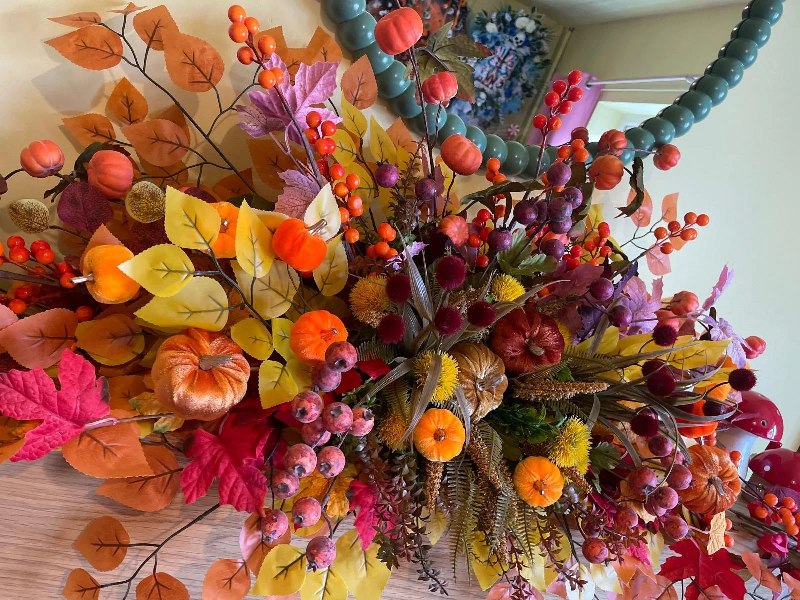 An autumn-themed floral arrangement featuring small pumpkins, berries, and colorful leaves in shades of orange, red, yellow, and purple on a wooden table.