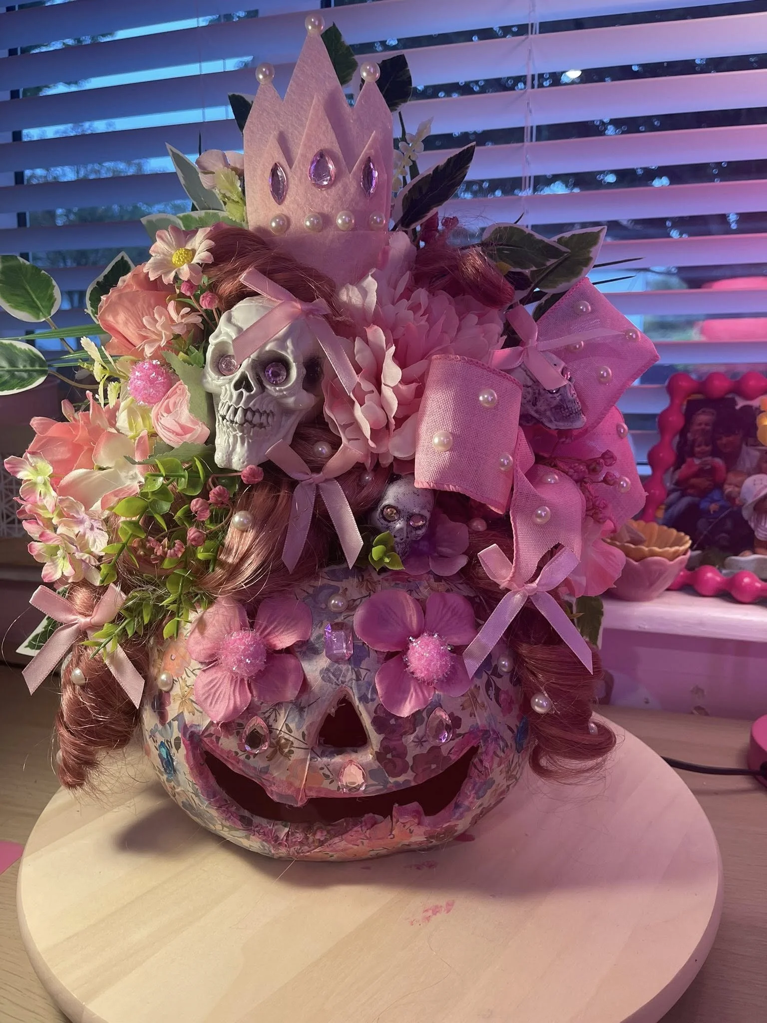 Decorated pumpkin with pink flowers, ribbons, small skulls, and a pink crown on top, set on a wooden round base in front of window blinds.
