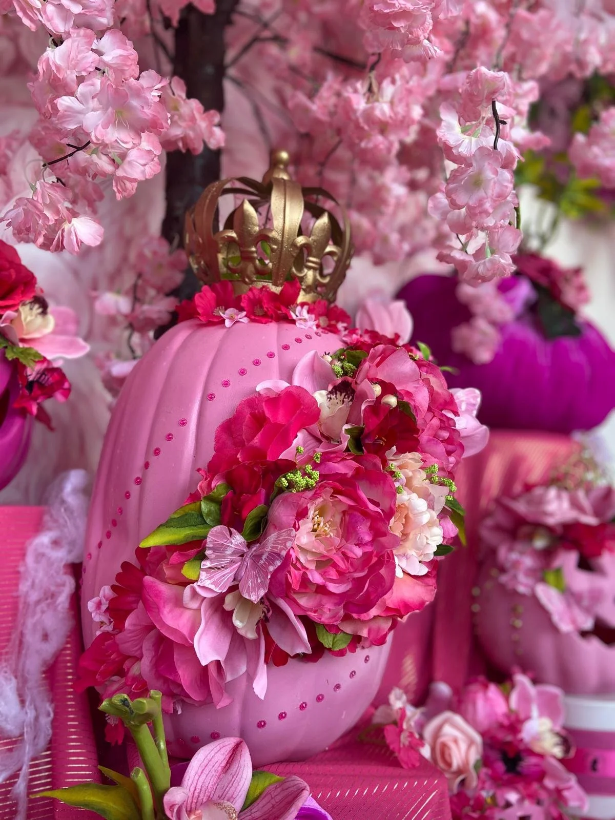 Pink decorative object shaped like an egg with pink and red flowers and green leaves, topped with a gold crown, surrounded by pink blossoms and flowers.