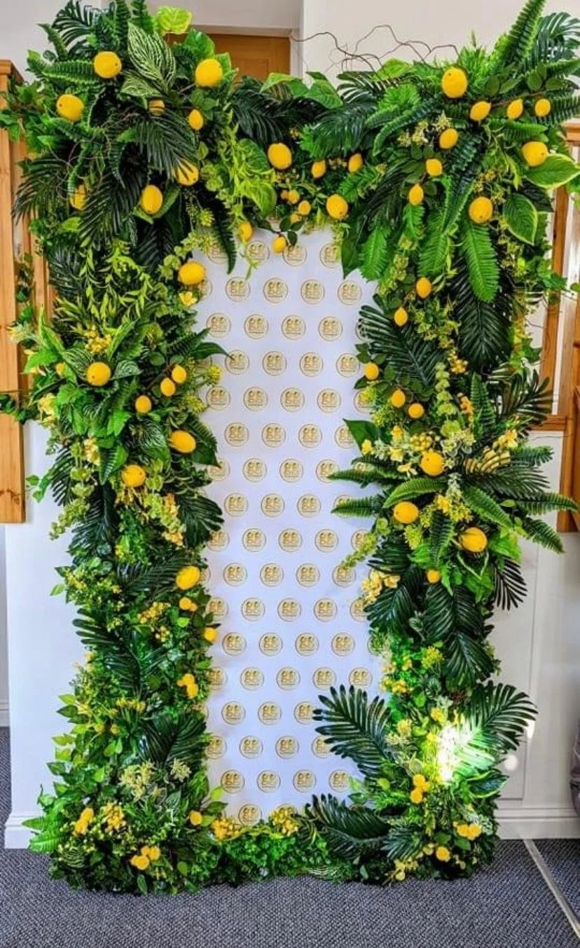 A decorative floral arrangement with lush green leaves and yellow accents, surrounding a branded backdrop with logos.