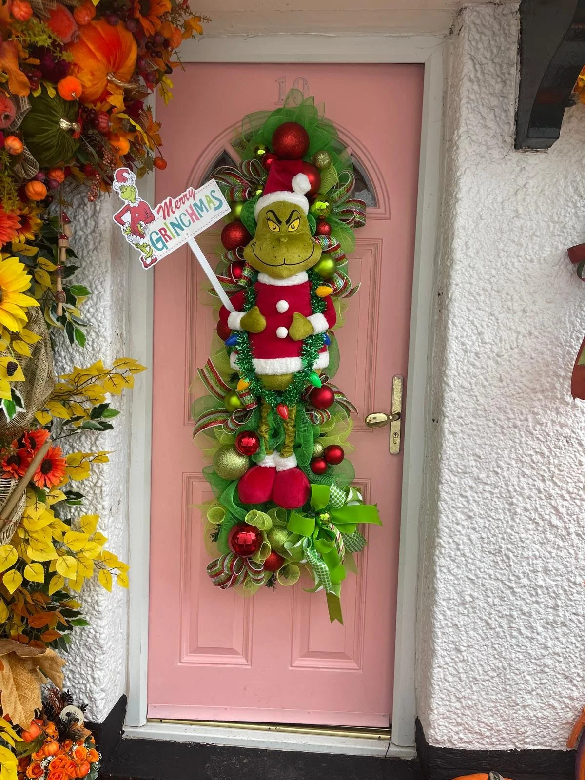Decorated pink front door with a large number 10 wreath featuring the Grinch wearing a Santa hat and red jacket, surrounded by red and green ornaments and ribbons, celebrating Christmas.