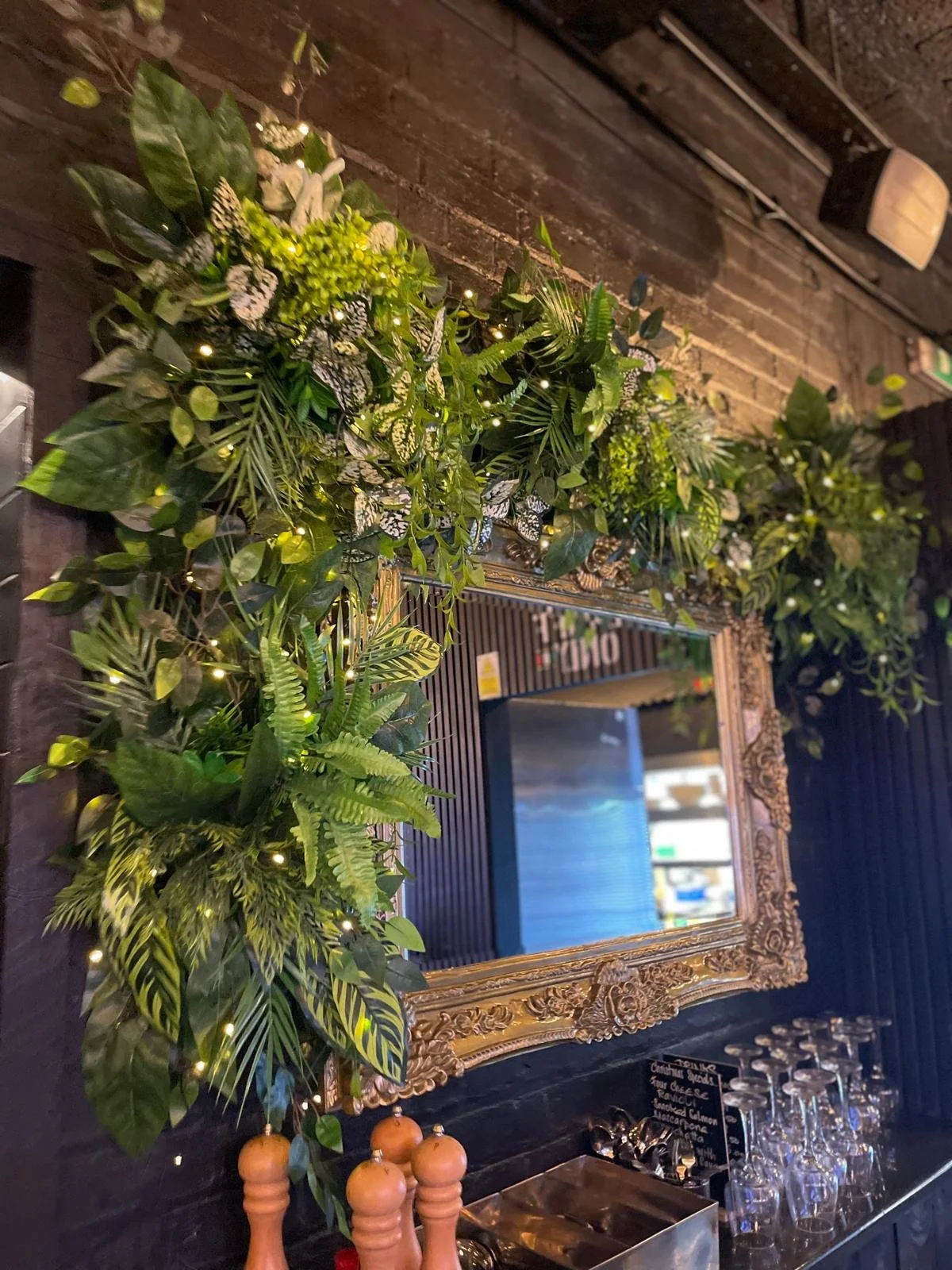 Decorative mirror surrounded by lush green foliage with fairy lights, mounted on a dark wooden wall, reflecting part of a bar or restaurant interior.