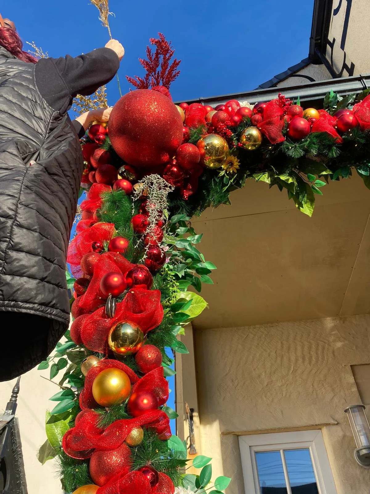 Close-up of holiday decoration with red, gold, and glittery ornaments, red mesh ribbons, green leaves, and berries on a building corner, with a person in a black jacket hanging a decoration.
