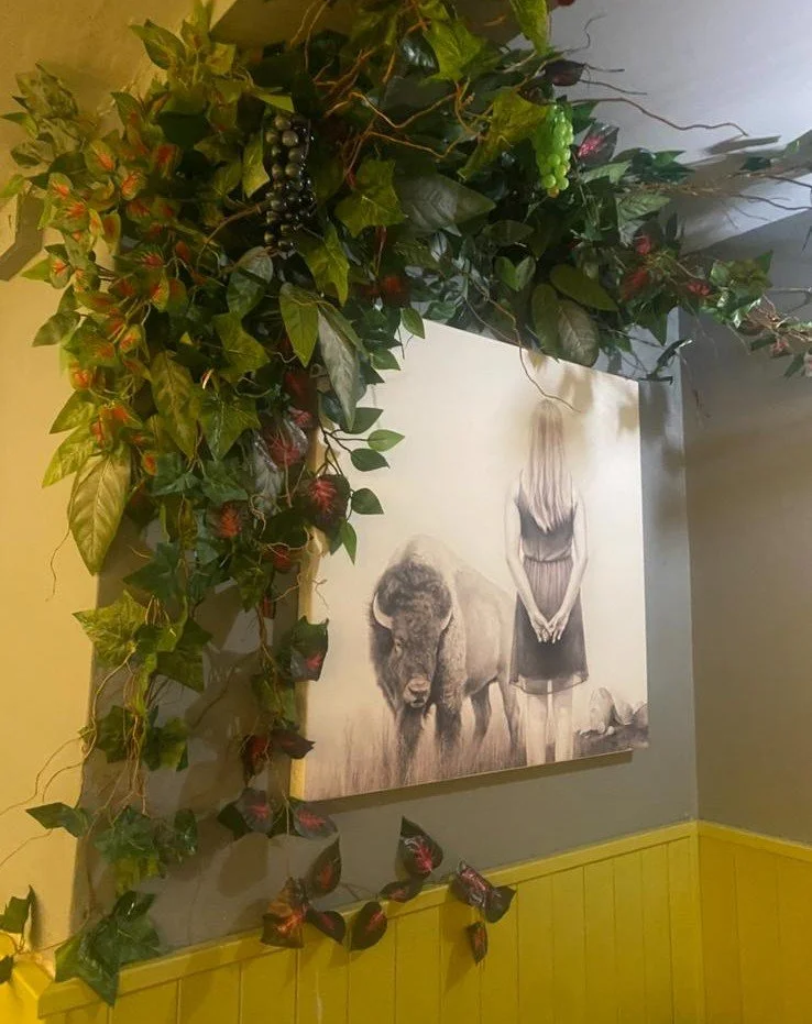 A painting of a woman with long blonde hair wearing a black dress, standing next to a buffalo, hangs on a wall decorated with green foliage and red berries.