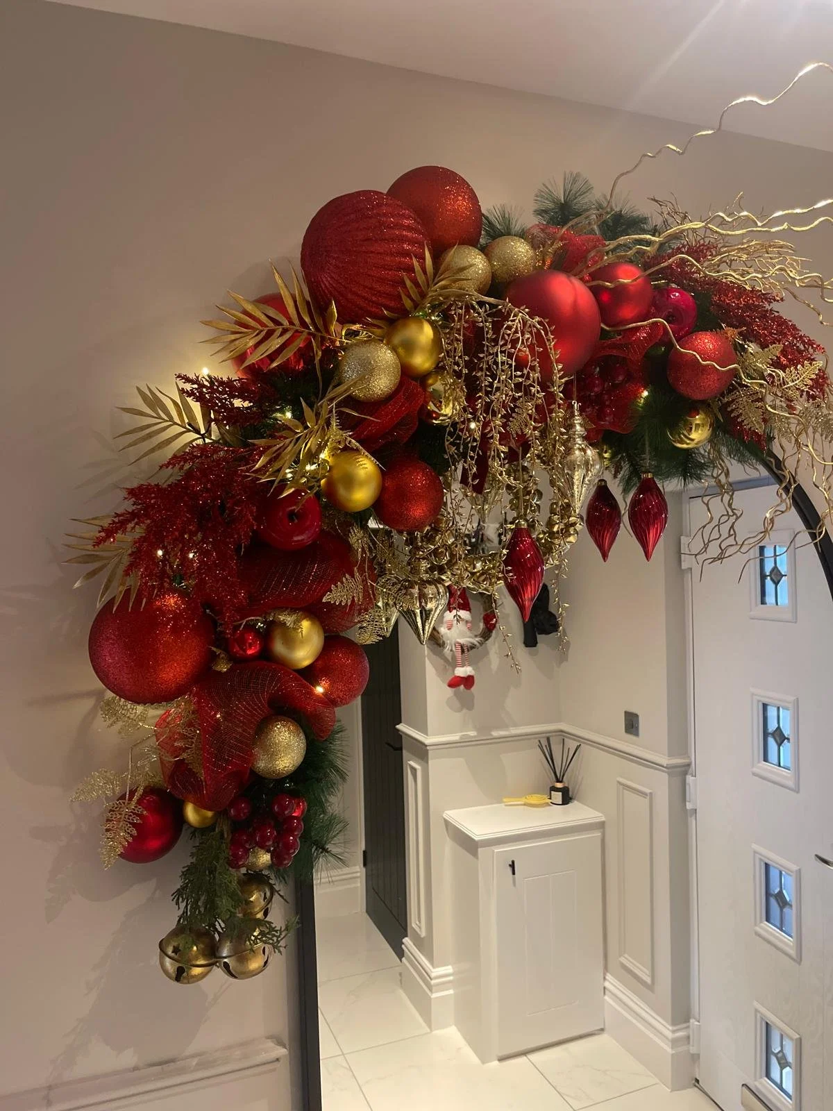 Large, festive Christmas wreath hanging above an interior doorway, decorated with red, gold, and glittery ornaments, gold leaves, gold branches, and string lights.
