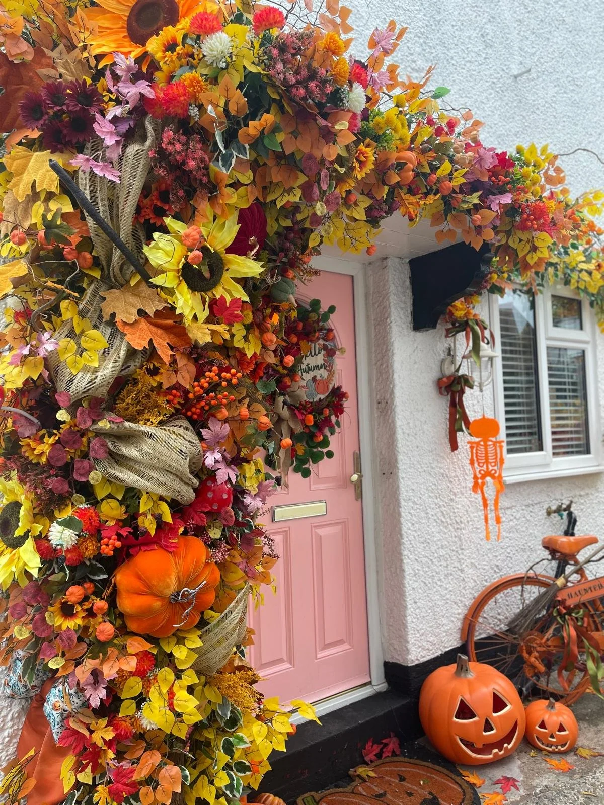 Decorated house entrance with fall-themed decorations: a pink door, a large rainbow-shaped fall wreath with sunflowers, pumpkins, autumn leaves, and berries; an orange pumpkin-shaped lantern, a small pumpkin decoration, and a decorated orange bicycle