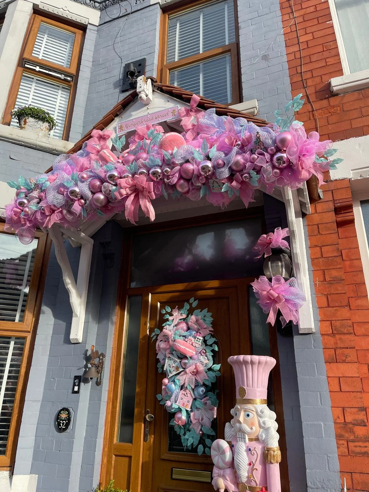 Decorated house entrance with pink holiday wreath, pink ornaments, ribbons, and bows. A large nutcracker statue dressed as a chef stands near the door.