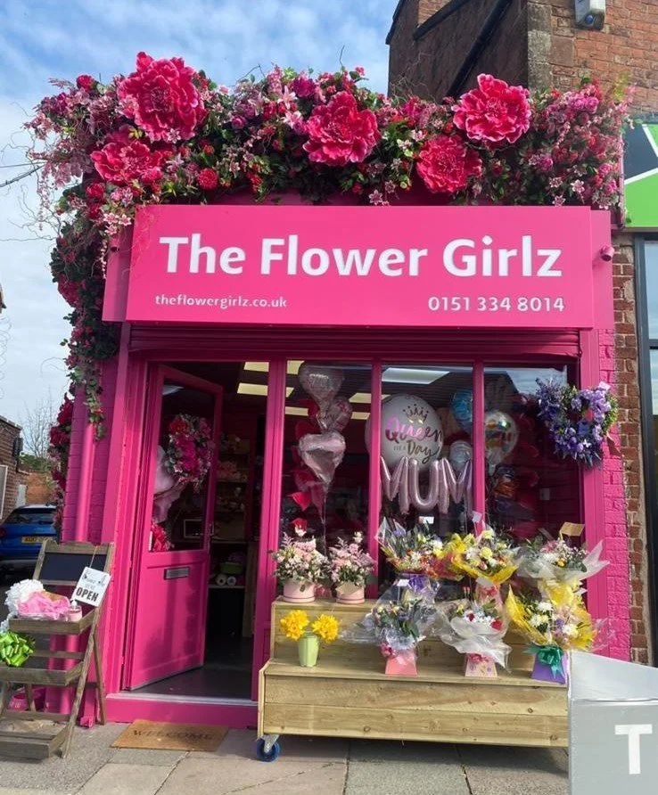 A bright pink flower shop called "The Flower Girlz" decorated with pink and purple flowers on the roof and outside. The shop has a display of colorful flower bouquets and balloons, including a "Queen Mum" balloon. There is a small wooden table with f