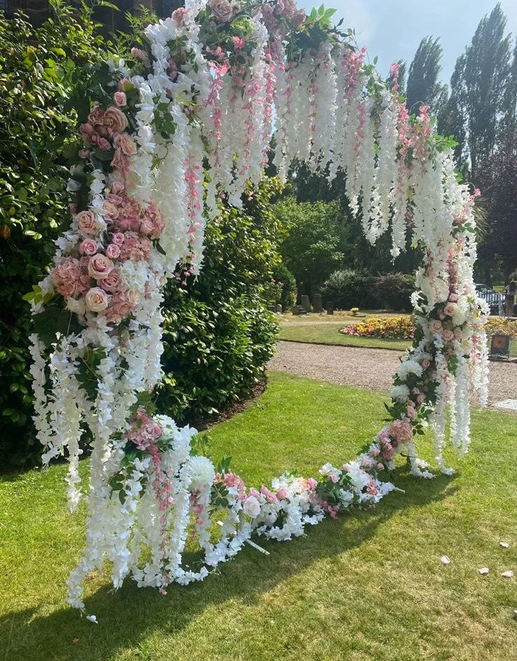 A circular floral arch decorated with pink and white roses and cascading white flowers, set on a grassy area in a garden or park.