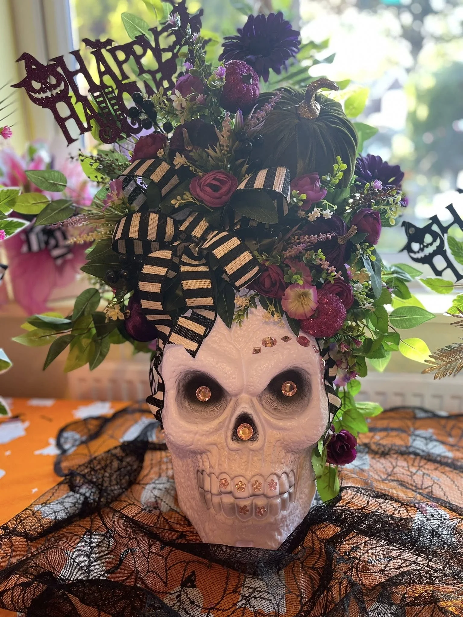 A Halloween-themed decorative skull with a floral arrangement on top, adorned with a black and white striped ribbon, rhinestones, and surrounded by black lace and spiderweb fabric.
