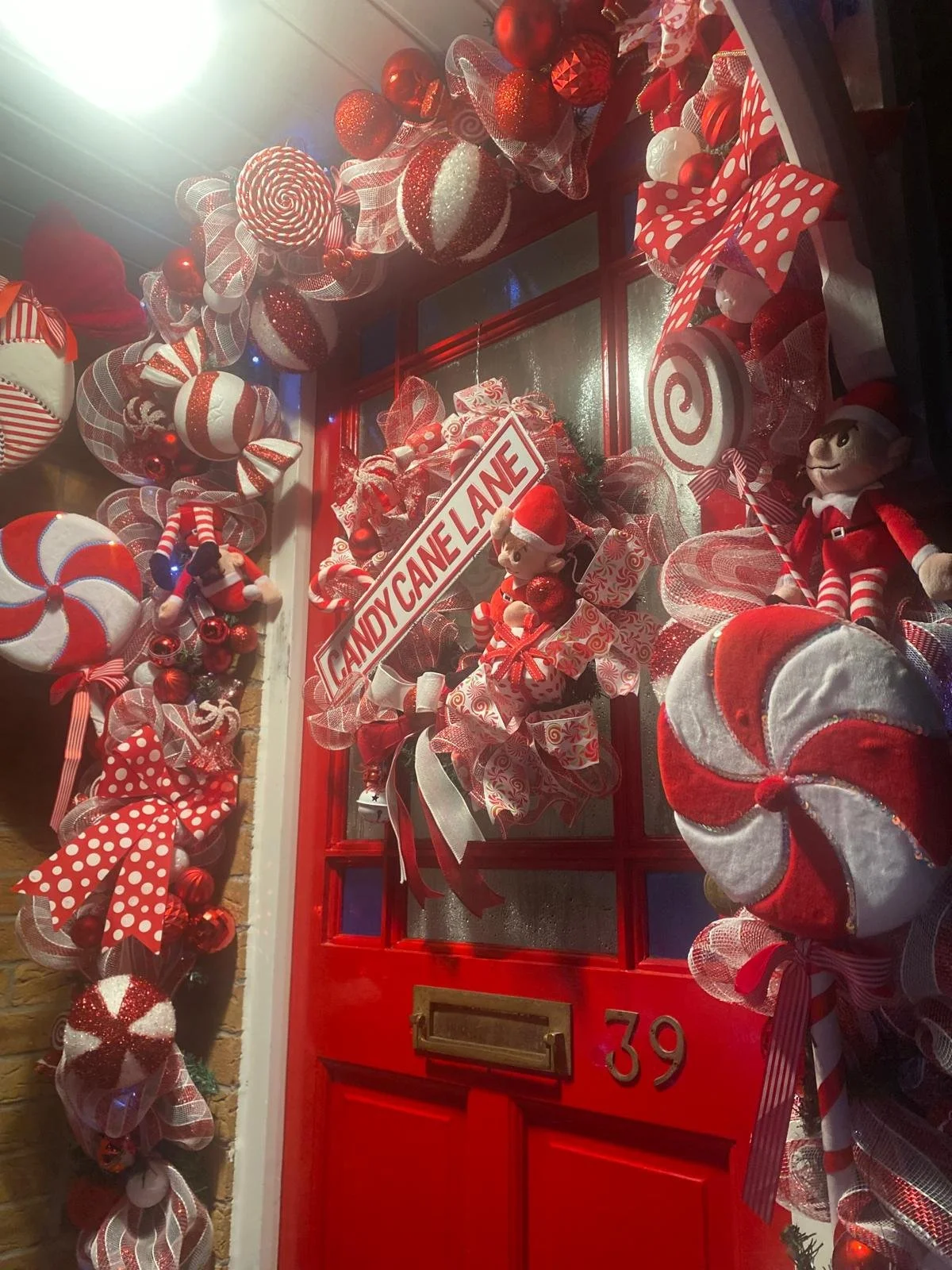 Christmas decorated red front door with holiday wreaths, ribbons, ornaments, and plush holiday characters, displaying a 'Candy Cane Lane' sign and the house number 39.