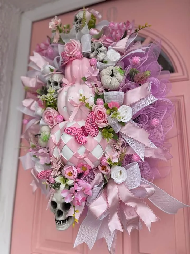 Decorative door wreath with pink, white, and purple ribbons, flowers, small skulls, pumpkins, and butterflies, hanging on a pink door.