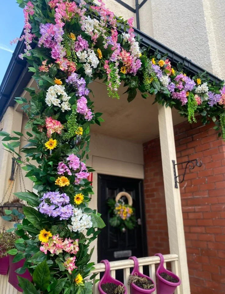 Decorative flower arch with pink, purple, white, and yellow flowers at a house entrance, with purple hanging planters filled with soil below.