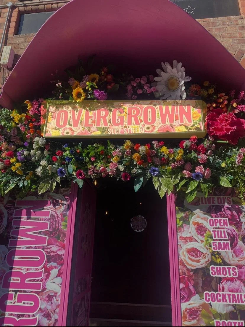 Colorful floral arrangement surrounding the entrance of a flower shop named 'Overgrown' on Self Street, with signage indicating it is open till 5AM and offers shots, cocktails, and other products.