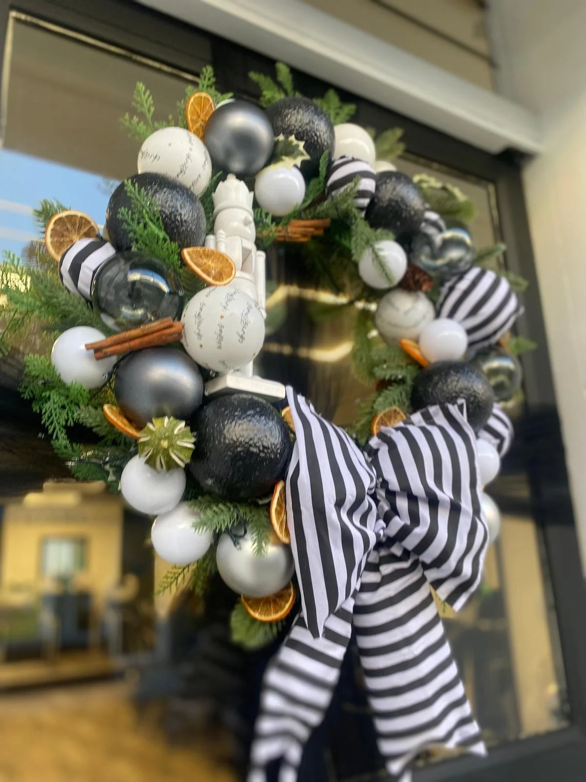 Christmas wreath with black, white, and silver ornaments, dried orange slices, cinnamon sticks, greenery, and a black and white striped bow hanging on a door.