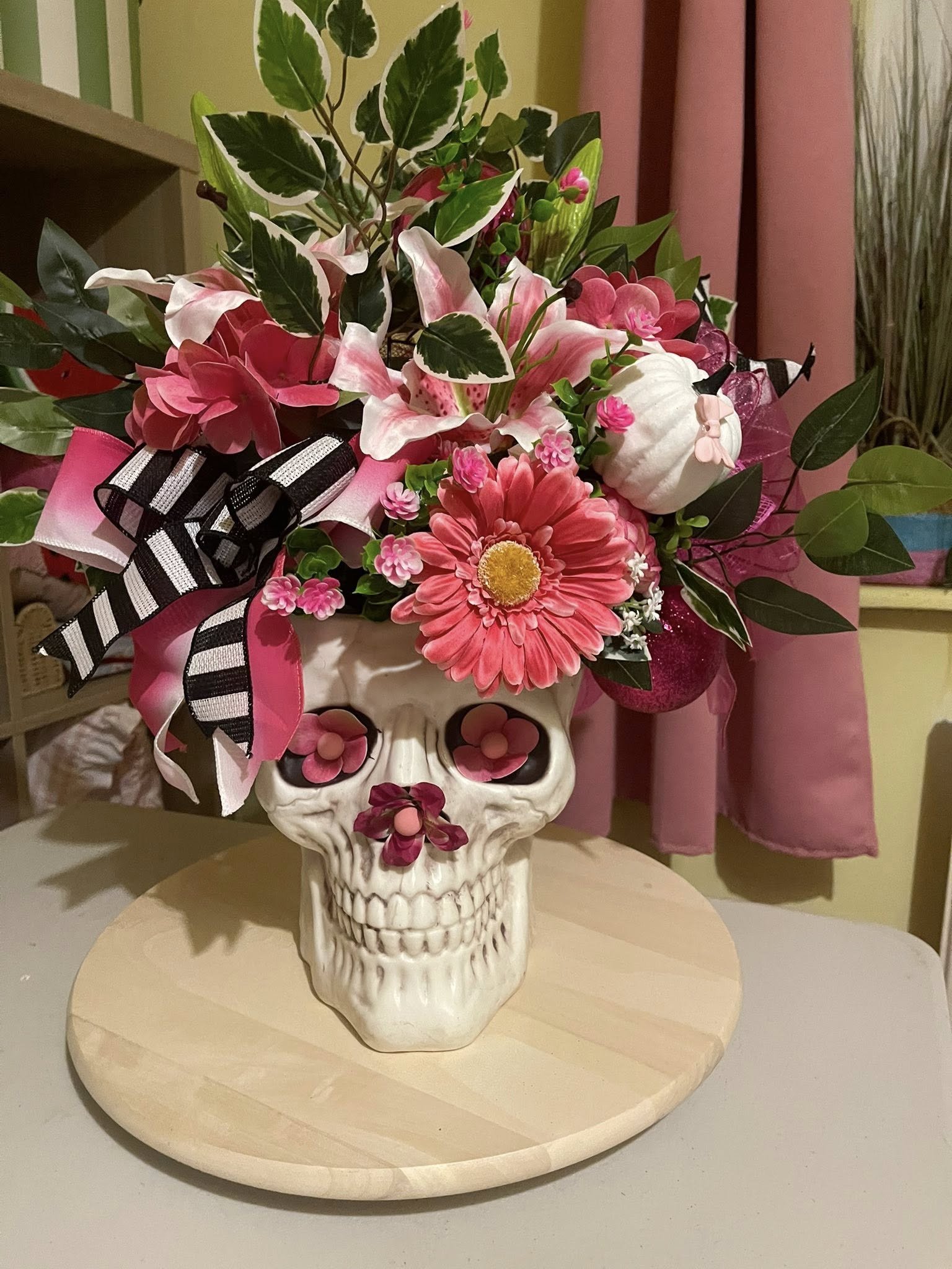 A decorative flower arrangement in a skull-shaped vase, featuring pink, white, and green flowers, with ribbons on the side, placed on a round wooden board.