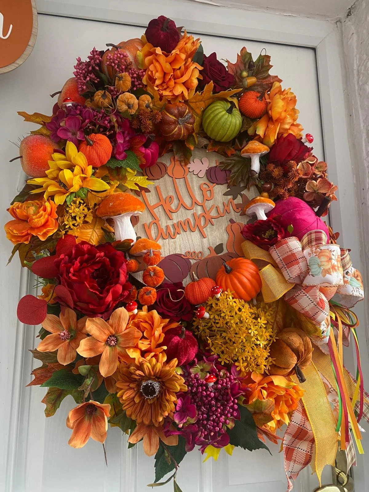 Fall-themed wreath made of artificial flowers, pumpkins, gourds, and ribbons hanging on a door with a wooden sign reading "Hello Pumpkin" in the center.