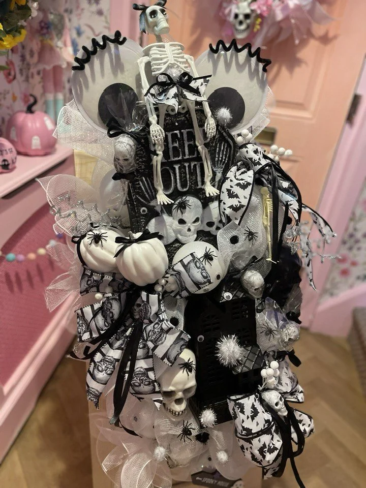 A Halloween-themed gift arrangement with skeletons, skulls, pumpkins, black bows, spiders, and bat decorations in black, white, and gray colors.