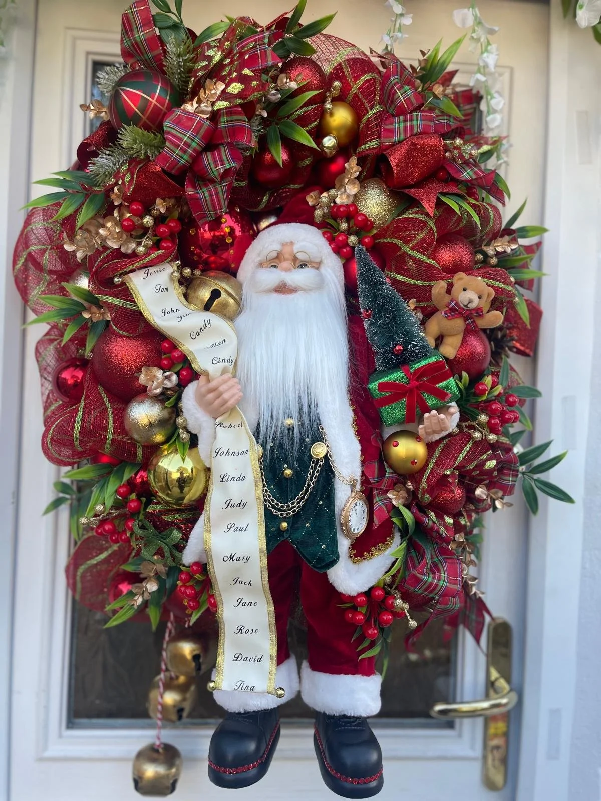 Large Christmas wreath decorated with red and gold ornaments, ribbons, and greenery. In the center is a Santa Claus figure holding a tiny Christmas tree and a list of names, wearing a traditional red and white outfit with a black belt and boots.