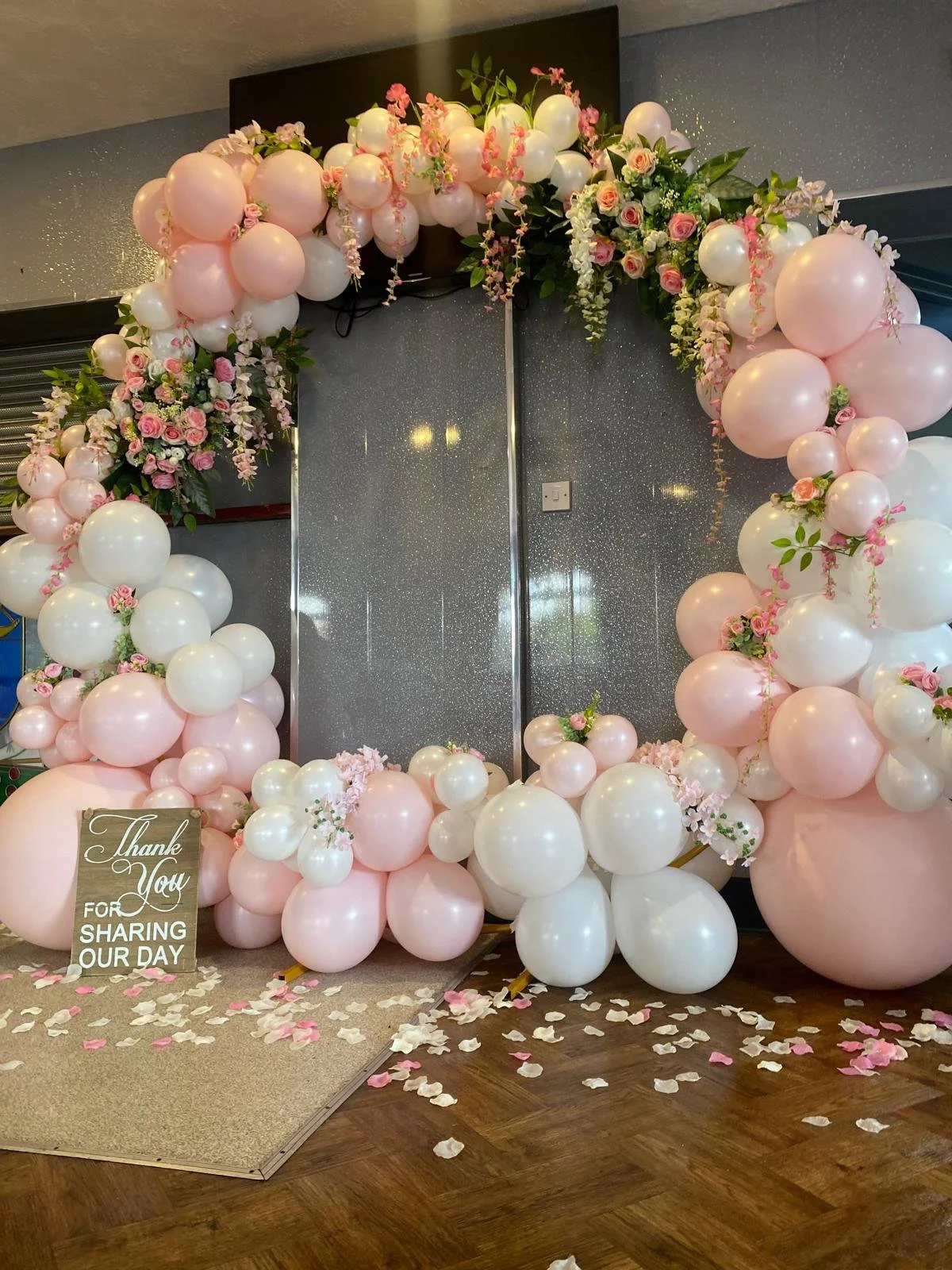 A balloon arch decoration with pink and white balloons, decorated with pink and white flowers, on a wall. There is a small sign that says 'Thank You for Sharing Our Day' surrounded by pink and white petals on the floor.