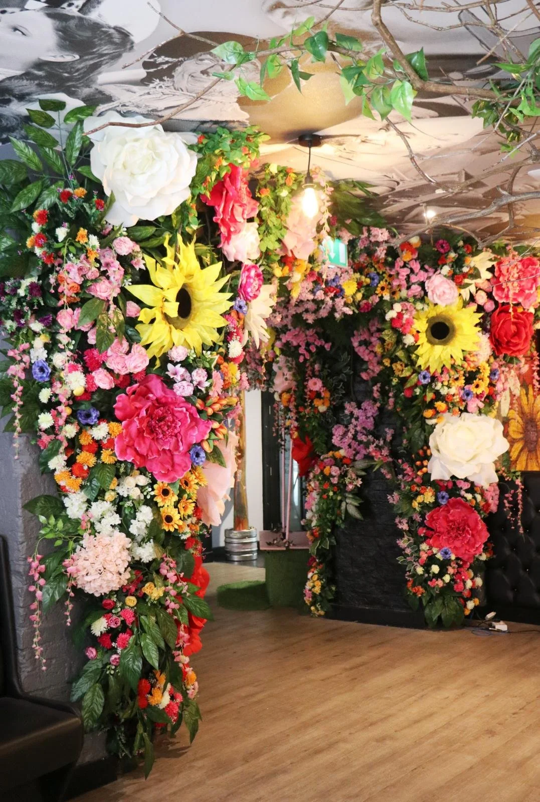 Colorful floral arch with pink, red, yellow, and white flowers, green leaves, and branches, set inside an indoor space with wooden floors and a mural on the ceiling.