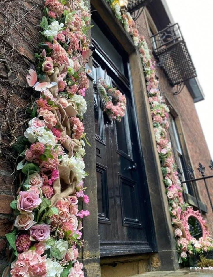A black front door decorated with pink and white floral arrangements, including roses and other flowers, along the door frame and on the ground.