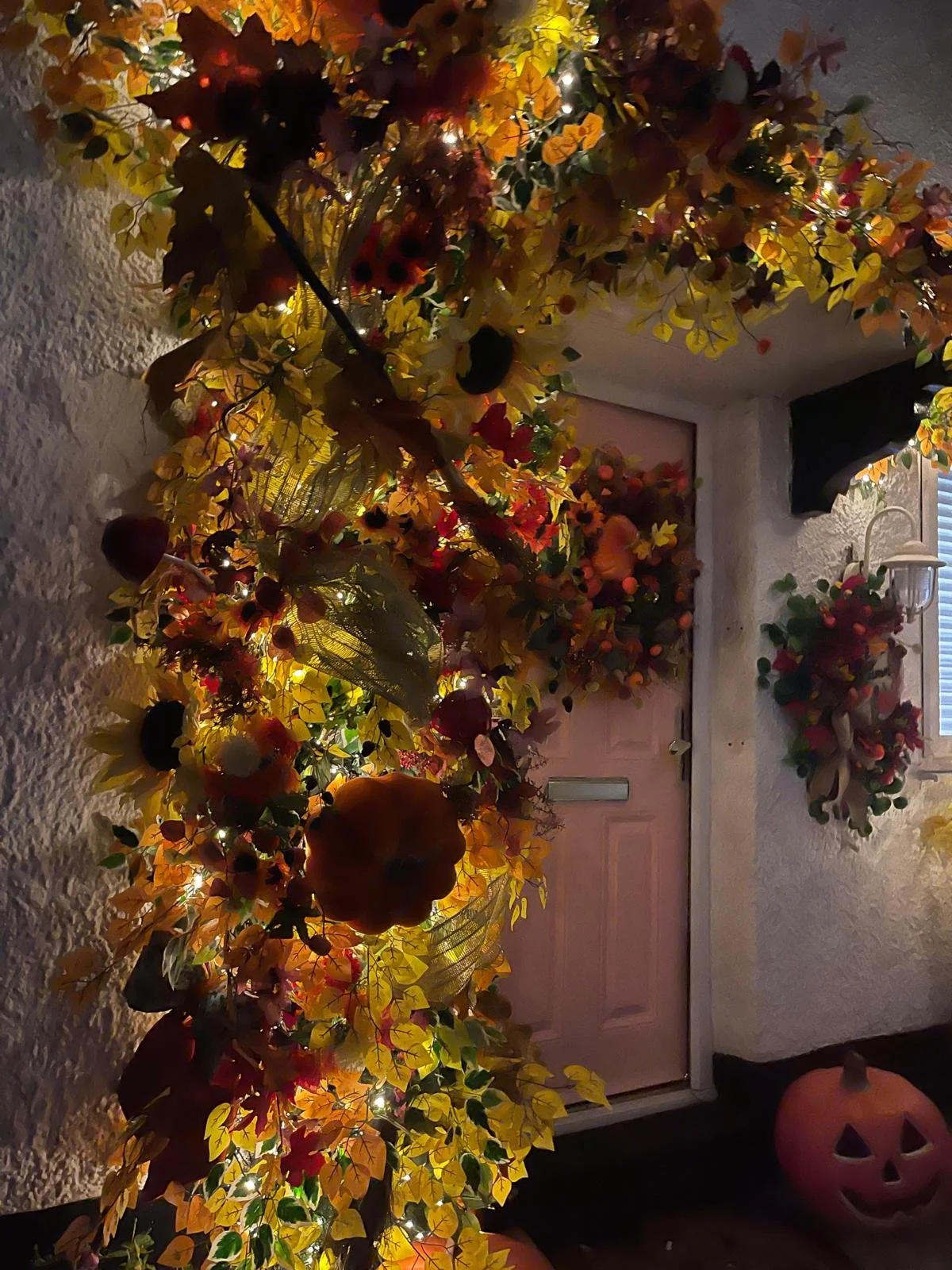 Decorative autumn-themed fall harvest display with artificial pumpkins and flowers, string lights, near a pink door, against textured white exterior wall.