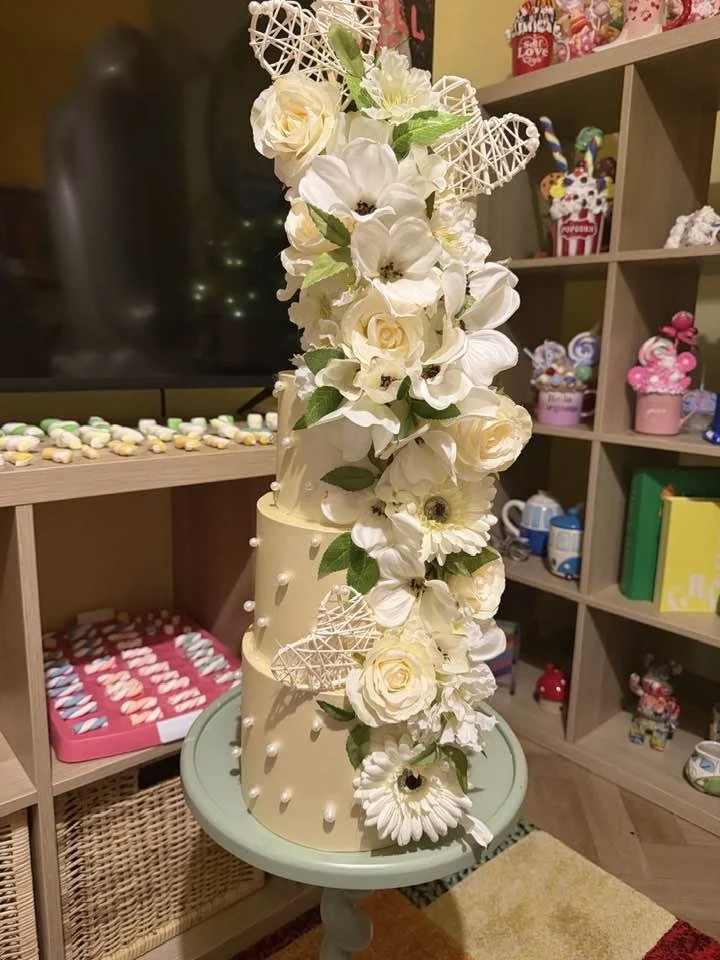 A two-tiered white wedding cake decorated with white flowers, green leaves, and decorative heart-shaped lattice accents, placed on a small round table.
