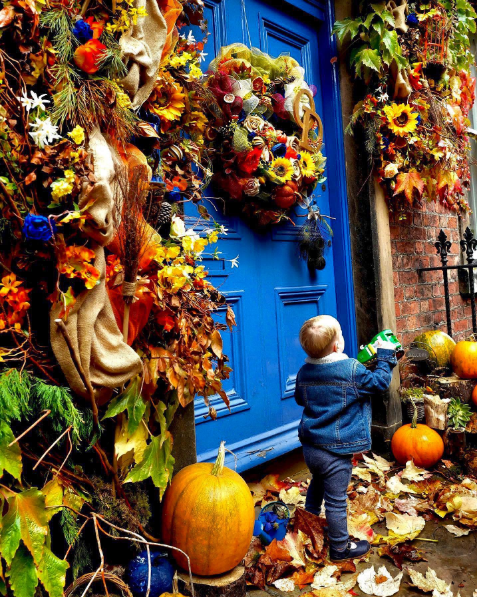 A young boy standing outside a house with a bright blue door, decorated with autumn-themed wreaths, pumpkins, and fall leaves.