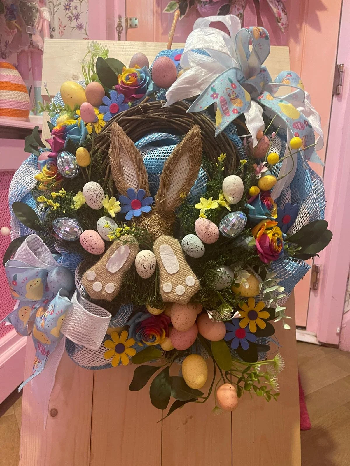 Easter basket decorated with colorful flowers, Easter eggs, bunny ears, bows, and ribbons, placed on a wooden table.