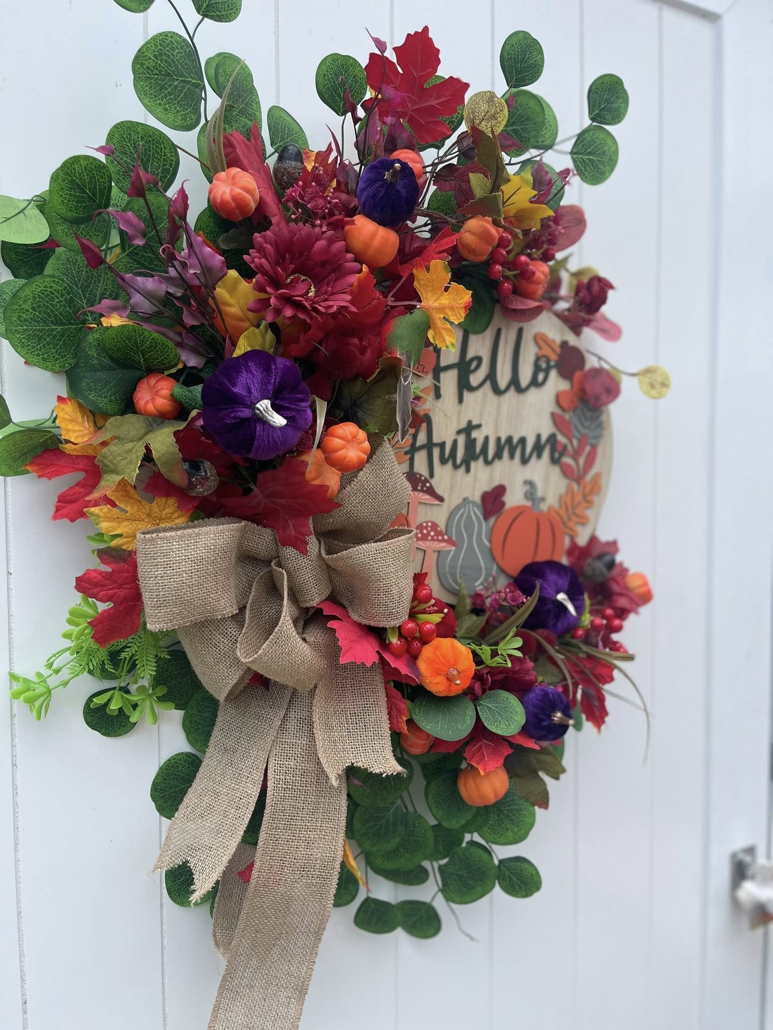 Fall-themed decorative wreath with colorful flowers, pumpkins, and a beige burlap bow, hanging on a white door with a sign that says "Hello Autumn".