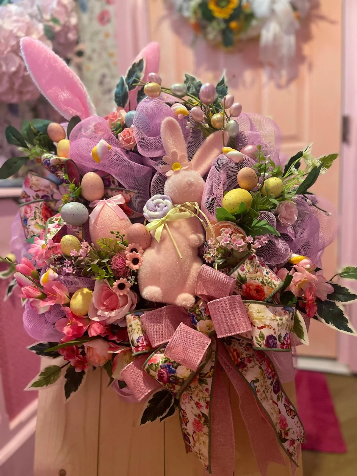A decorative arrangement of pastel-colored Easter-themed items, including a plush bunny, decorated eggs, flowers, ribbons, and faux foliage, all set against a pink background.