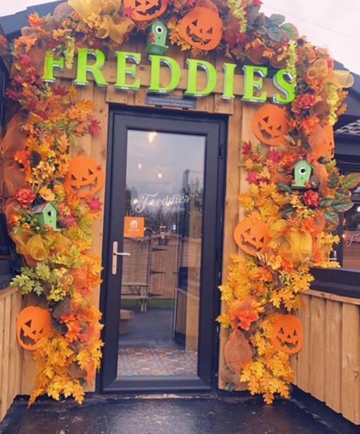 Decorative entrance with autumn leaves and Halloween pumpkin decorations around a door, featuring a sign that says "FREDDIES" in bright green letters.