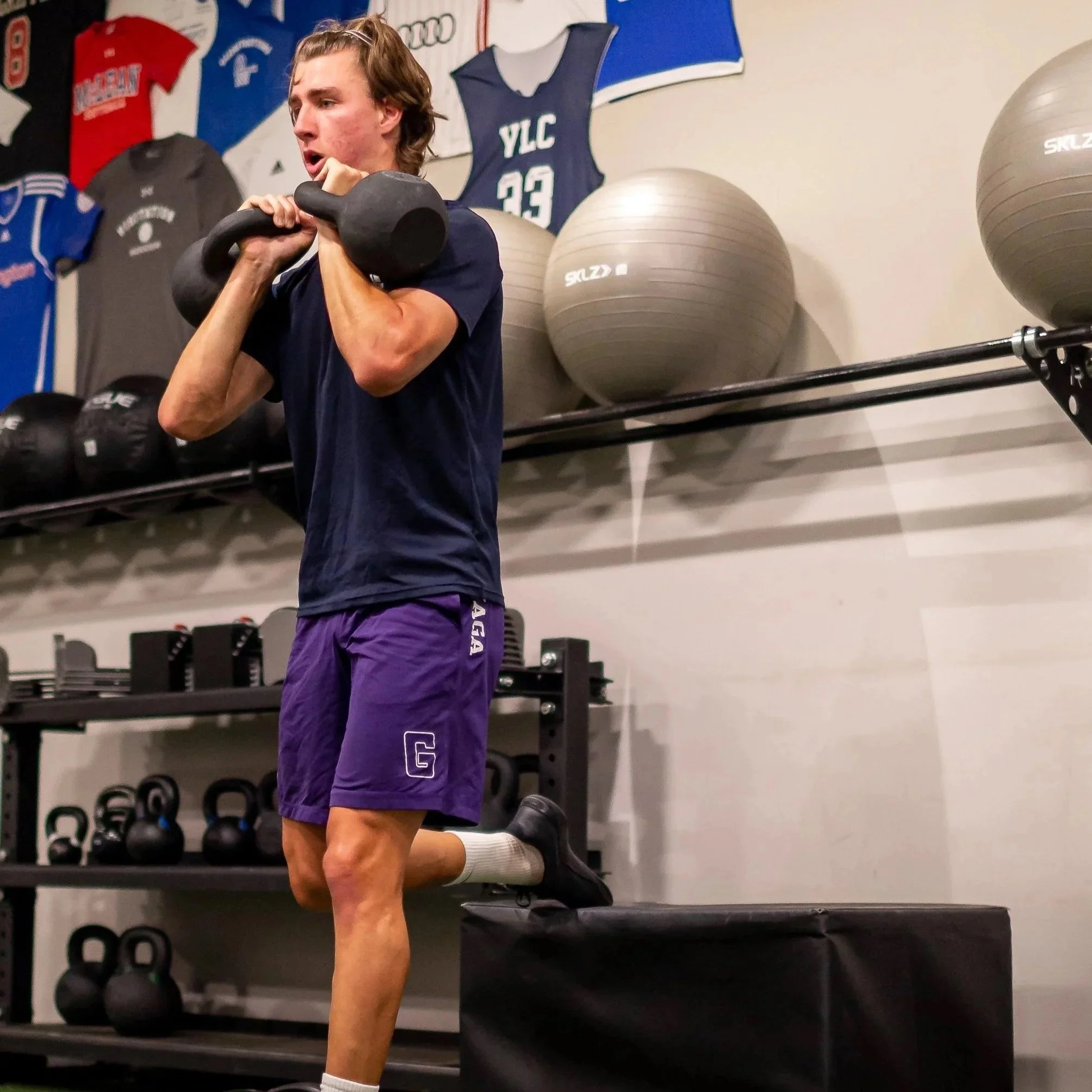 A young man in a gym performing a kettlebell exercise, surrounded by exercise equipment and sports jerseys on the wall.