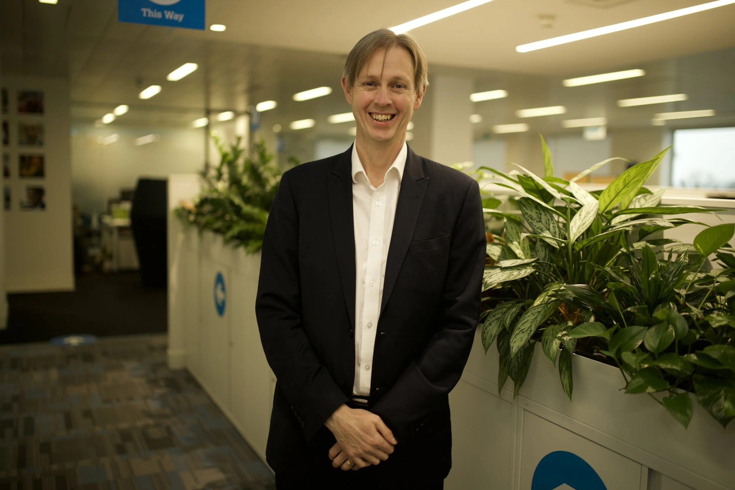 A man in a dark suit and white shirt standing in an office with green plants and fluorescent lighting, smiling at the camera.