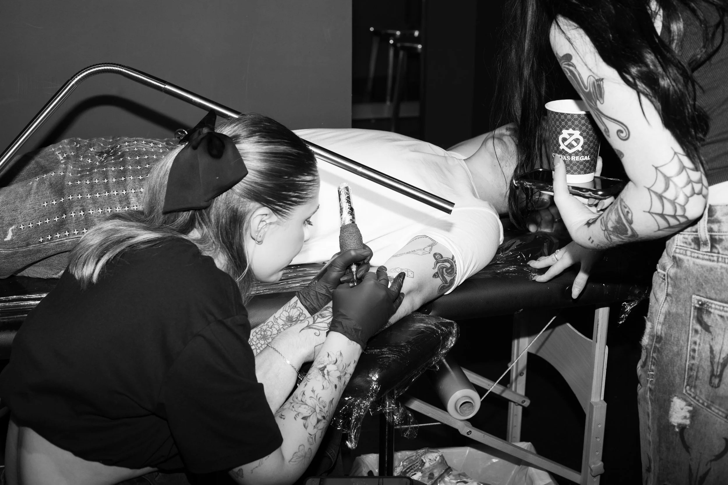 Tattoo artist working on a client's arm while another person holds a coffee cup nearby