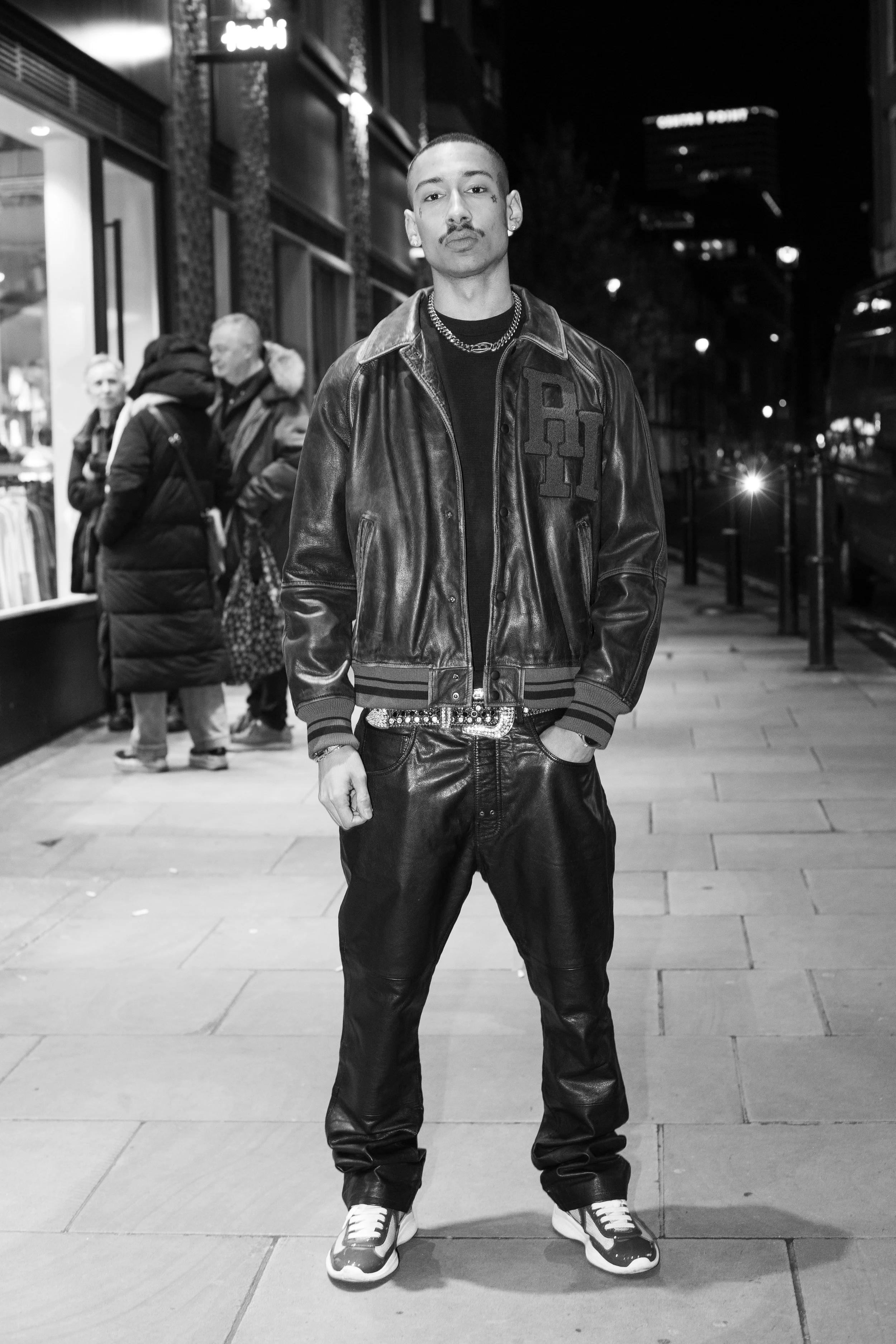 A man standing on a city sidewalk at night, wearing a leather jacket, chain necklaces, and sneakers, with a group of people in the background near a storefront.